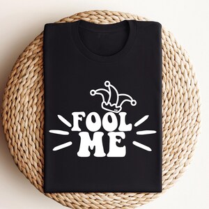 Puede incluir: Camiseta negra con el texto blanco "Fool Me" y un gr&aacute;fico de sombrero de buf&oacute;n. La camiseta est&aacute; doblada sobre una superficie circular tejida. El dise&ntilde;o utiliza una fuente retro y juguetona.