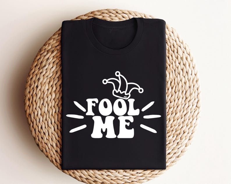 Puede incluir: Camiseta negra con el texto blanco "Fool Me" y un gr&aacute;fico de sombrero de buf&oacute;n. La camiseta est&aacute; doblada sobre una superficie circular tejida. El dise&ntilde;o utiliza una fuente retro y juguetona.