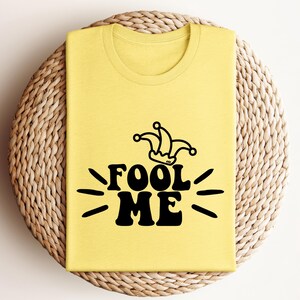 Puede incluir: Una camiseta amarilla clara doblada con la frase negra "FOOL ME" y un dise&ntilde;o de sombrero de buf&oacute;n negro encima. La camiseta est&aacute; colocada sobre una superficie redonda tejida. El dise&ntilde;o est&aacute; en una fuente retro audaz.