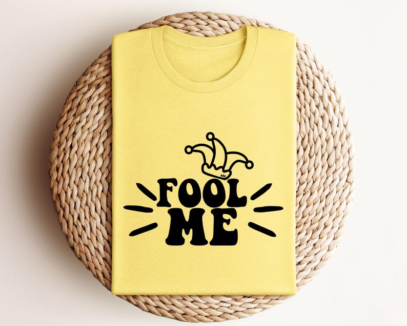 Puede incluir: Una camiseta amarilla clara doblada con la frase negra "FOOL ME" y un dise&ntilde;o de sombrero de buf&oacute;n negro encima. La camiseta est&aacute; colocada sobre una superficie redonda tejida. El dise&ntilde;o est&aacute; en una fuente retro audaz.