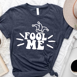 Puede incluir: Camiseta azul marino oscuro con la frase blanca "FOOL ME" y un gr&aacute;fico de sombrero de buf&oacute;n. La camiseta tiene cuello redondo. Un sombrero y una taza de caf&eacute; son visibles en el fondo.