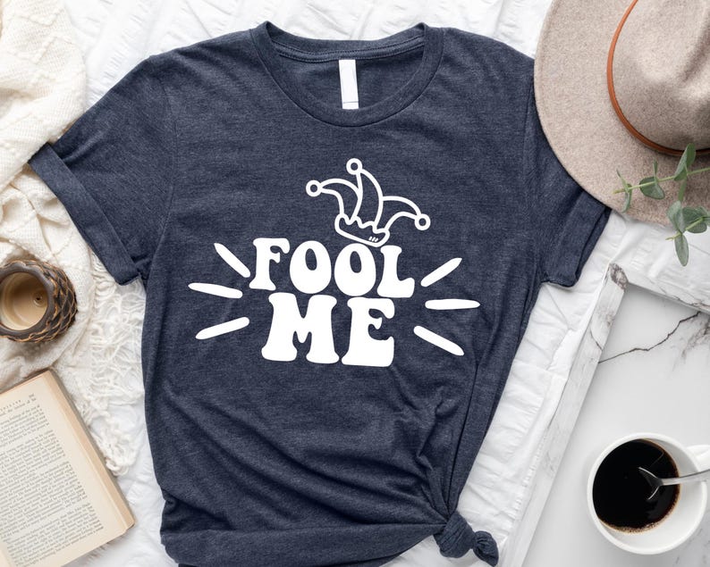 Puede incluir: Camiseta azul marino oscuro con la frase blanca "FOOL ME" y un gr&aacute;fico de sombrero de buf&oacute;n. La camiseta tiene cuello redondo. Un sombrero y una taza de caf&eacute; son visibles en el fondo.
