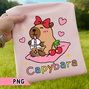 Peut inclure: T-shirt rose clair avec un dessin de capybara de dessin animé. Le capybara porte un nœud rouge et boit du thé aux perles. Le motif comprend des cerises, des cœurs et le mot "Capybara".