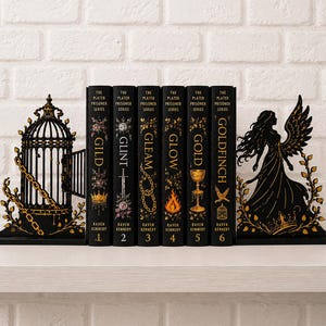 Può includere: Un set di sei libri neri della serie The Plated Prisoner di Raven Kennedy, intitolati Gild, Glint, Gleam, Glow, Gold e Goldfinch, posizionati tra due fermalibri in metallo nero con un design a gabbia per uccelli e una figura alata.