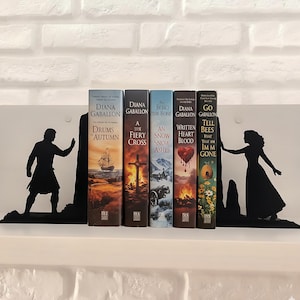 Peut inclure: Des serre-livres en métal noir avec des silhouettes d'un homme et d'une femme, soutenant une rangée de livres. Les livres ont des couvertures colorées et des titres de Diana Gabaldon. Les serre-livres sont sur une étagère blanche contre un mur de briques blanches.