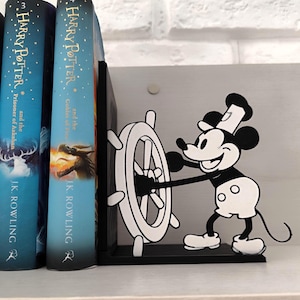 Pode incluir: Um suporte de livros temático do Mickey Mouse em preto e branco com Mickey Mouse segurando um leme. O suporte de livros é colocado ao lado de vários livros de Harry Potter em uma prateleira branca, contra uma parede de tijolos brancos.