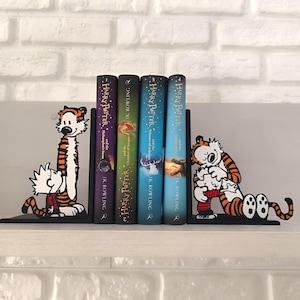 Pode incluir: Suportes de livros de metal preto com personagens de desenhos animados em laranja e preto. Seguram livros de Harry Potter. Os suportes de livros estão em uma prateleira branca contra uma parede de tijolos brancos.