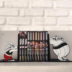 Op de afbeelding: Zwarte metalen boekensteunen met stripfiguren, die een rij manga-boeken vasthouden. De boeken zijn zwart met kleurrijke covers en de tekst "GACHIAKITA" en nummers. De boekensteunen staan op een witte plank tegen een witte bakstenen muur.