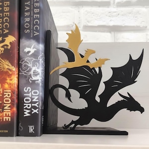 Pode incluir: Suportes de livros de dragão de metal preto com um detalhe de dragão dourado. Os suportes de livros são projetados para segurar livros e são colocados em uma prateleira branca. Os livros têm títulos como "Iron Flame" e "Onyx Storm".
