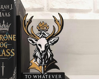 Könnte beinhalten: Schwarzer Buchstütze mit einem stilisierten Hirschkopf-Design mit Geweih, Krone und Schwert. Der Hirsch ist weiß, gold und schwarz. Auf der Basis des Buchstütze steht der Text "TO WHATEVER END". Links ist ein Buch mit goldfarbenen Buchstaben.