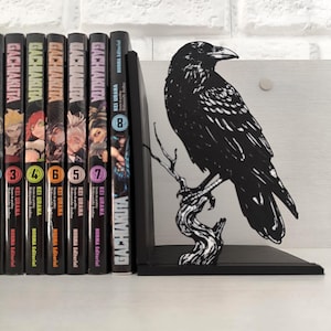 May include: A black raven-shaped bookend, perched on a branch, stands next to a row of colorful manga books. The bookend is made of a solid material and is designed to hold books upright on a shelf. The books are numbered and titled "Gachakita".