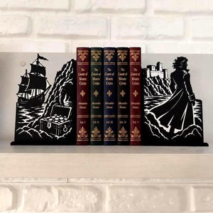 Op de afbeelding: Een set zwarte metalen boekensteunen met een silhouet van een zeilschip en een schatkist aan de linkerkant, en een figuur voor een kasteel aan de rechterkant, die vijf delen van De Graaf van Monte-Cristo van Alexandre Dumas vasthouden.
