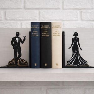 Może przedstawiać: Czarne metalowe podpórki do książek z sylwetkami w stylu art deco mężczyzny i kobiety. Podpórki do książek flankują trzy egzemplarze "The Great Gatsby" F. Scotta Fitzgeralda. Książki są granatowe, czarne i kremowe.
