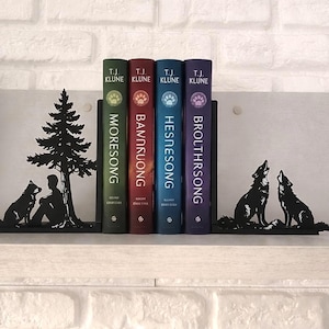 Könnte beinhalten: Vier Bücher mit farbenfrohen Buchrücken, gehalten von schwarzen Metall-Buchstützen. Die Buchstützen zeigen Silhouetten von Wölfen und einer Person mit einem Hund unter einem Baum. Die Buchtitel lauten "MORESONG", "BANNKUONG", "HESNESONG" und "BROUTHRSONG".