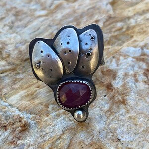 May include: A silver ring featuring a cluster of three textured, silver, organic shapes with a deep red gemstone at its centre.