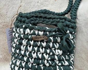 May include: A dark green and white crocheted handbag featuring a thick braided shoulder strap and a small decorative bow on the side. A brown leather-like tag is attached to the front with the text Fadenzauber and two small heart symbols.