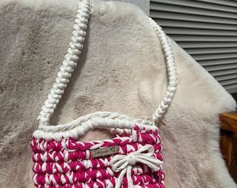 May include: A handmade crochet handbag featuring a vibrant magenta and white colour pattern. The bag includes a thick braided white shoulder strap, a white crochet bow detail on the front, and a small rectangular wooden label with the text Fadenzauber.