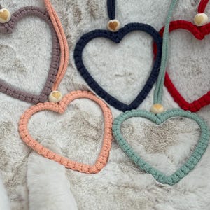 以下が含まれることがあります： ピンク、青、赤、緑など、さまざまな色の手作りのマクラメハートオーナメントのコレクション。各ハートは、木製のビーズが付いたコードに取り付けられています。柔らかく、質感のある表面に飾られています。