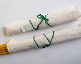 May include: Two bundles of yellow beeswax candles wrapped in white fabric and tied with green twine. The fabric has a green label with the text "HANDMADE SOY & BEESWAX CANDLES".