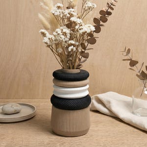 Puede incluir: Un jarrón beige apilado con anillos negros y blancos contiene flores secas. También hay un pequeño jarrón de vidrio con eucalipto y un plato pequeño, redondo y beige con una piedra. Sobre una superficie de madera.