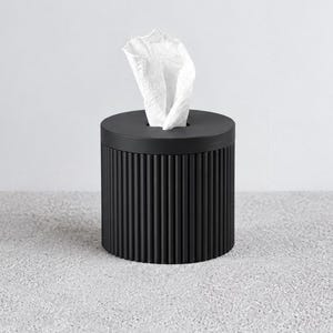 Puede incluir: Un dispensador de pañuelos cilíndrico de color negro con una textura acanalada vertical en el exterior. La superficie superior es plana con una abertura circular central, a través de la cual sobresale un pañuelo blanco hacia arriba.