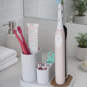 May include: A grey toothbrush organiser with a textured finish, holding toothbrushes, toothpaste, and floss picks. Two electric toothbrushes stand on a brown, textured base. The bathroom accessory is on a white countertop.