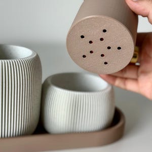 May include: A set of three small, neutral-toned planters. One is held up, revealing drainage holes. The other two are in a tray. The planters have vertical ribbed detailing. The colours are beige, white, and brown.