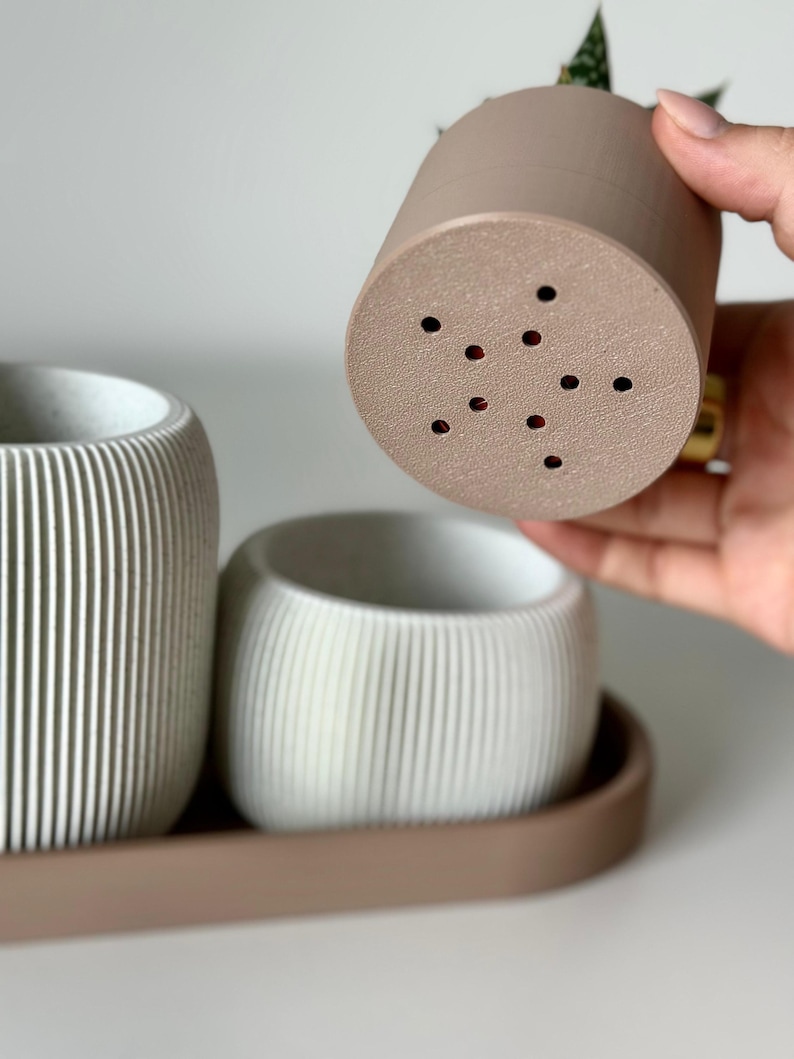 May include: A set of three small, neutral-toned planters. One is held up, revealing drainage holes. The other two are in a tray. The planters have vertical ribbed detailing. The colours are beige, white, and brown.