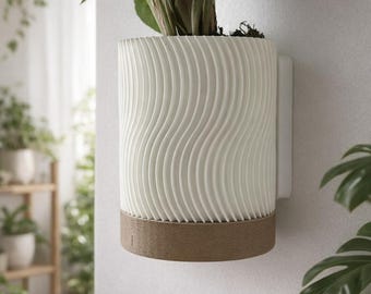 Könnte beinhalten: Ein weißer, zylindrischer Wandtopf mit einem wellenförmigen, strukturierten Design. Der Topf hat einen braunen Sockel und ist an einer weißen Halterung befestigt. Eine grüne Pflanze wächst im Topf und verleiht der Dekoration einen Hauch von Natur.
