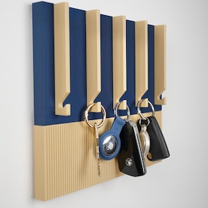 Può includere: Un portachiavi blu e beige dal design moderno. Il portachiavi ha ganci verticali beige e uno sfondo blu. Diverse chiavi e portachiavi sono appesi ai ganci.