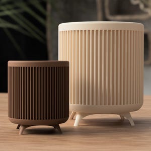 Op de afbeelding: Twee cilindrische plantenbakken, één bruin en één crème, met verticale geribbelde details. Beide plantenbakken hebben drie kleine, schuine poten. De plantenbakken worden getoond op een lichtgekleurd houten oppervlak, met een vage achtergrond van groen.