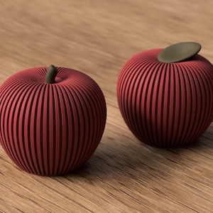 Op de afbeelding: Twee rode, appelvormige decoratieve objecten met een opvallende verticale geribbelde textuur. Elke appel heeft een glad, donkerbruin steeltje of blaadje in het midden aan de bovenkant. De objecten zijn afgerond en vertonen een symmetrische vorm.