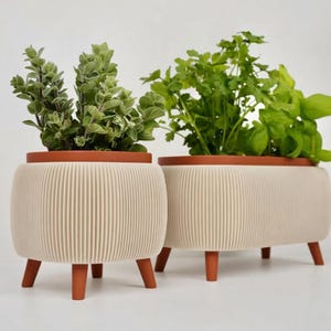 May include: Two cream-coloured planters with ribbed sides and terracotta rims and legs. Each planter holds a different type of green herb. The planters are designed with a modern aesthetic, suitable for indoor or outdoor use.