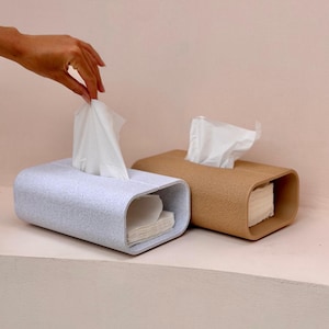 Peut inclure: Deux boîtes à mouchoirs rectangulaires aux coins arrondis. L'une est gris clair avec une texture mouchetée, et l'autre est beige avec une surface texturée. Chaque boîte contient une pile de mouchoirs en papier blancs, un mouchoir est tiré.