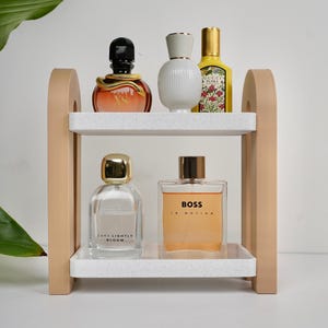 Może przedstawiać: Dwupoziomowa półka na perfumy w kolorze beżowo-białym, prezentująca różne butelki perfum. Półka ma nakrapianą białą powierzchnię i beżowe podpory. Perfumy obejmują butelki ze złotymi, czarnymi i białymi zakrętkami. Jedna butelka ma napis "BOSS IN MOTION".