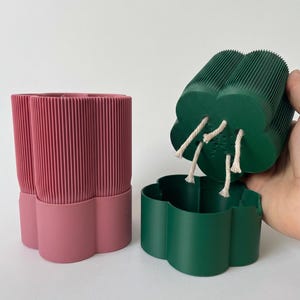 Può includere: Un set di due candele decorative. Una è una candela a forma di fiore di colore verde scuro, con quattro stoppini. L'altra è una candela rosa con un design a coste. Le candele sono di cera e sono decorative.