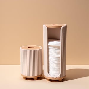 Könnte beinhalten: Zwei zylindrische Organizer mit gerippter hellrosa Außenseite sowie Ober- und Unterseite aus hellem Holz. Der höhere Organizer hat einen vertikalen Ausschnitt, der einen Stapel weißer, runder Wattepads zeigt, während der kürzere geschlossen ist.