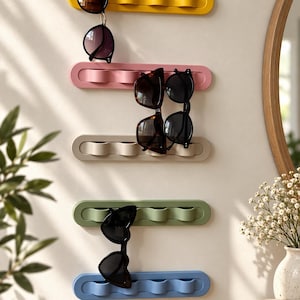 May include: Five horizontal wall-mounted organisers in yellow, pink, beige, green, and blue. Each organiser features four rounded, wave-like slots designed to hold items such as sunglasses. Several pairs of sunglasses are hanging from the slots.