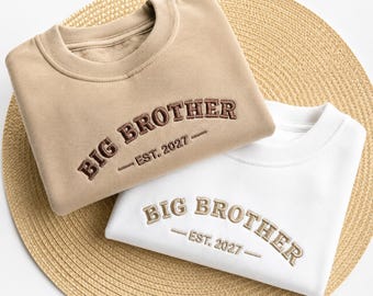 May include: A tan sweatshirt with dark brown embroidered text reading BIG BROTHER EST. 2027 and a white sweatshirt with tan embroidered text reading BIG BROTHER EST. 2027. Both garments feature a crew neckline and long sleeves in a folded arrangement.