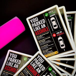 Op de afbeelding: Een stapel rechthoekige kaarten met een zwarte achtergrond en witte tekst "YOU PARKED LIKE AN ASSHOLE" in grote letters, naast een fel neonroze rechthoekig doosje. De kaarten bevatten diagrammen die correct en fout parkeren vergelijken.