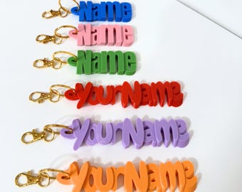 May include: Six colorful keychains with the words "Name" and "Your Name" in a 3D font. The keychains are made of plastic and have a gold metal ring.