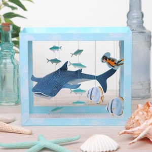 Peut inclure: Un diorama en papier bleu représentant un requin-baleine, divers poissons et un plongeur. La scène est encadrée dans un cadre bleu clair, avec les créatures marines suspendues par des fils. Le fond comprend des coquillages et une étoile de mer.