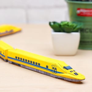May include: A yellow toy train with blue and black details. The train is on a wooden surface, with a small white pot and a green plant in the background. The train has a detailed design.