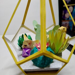 May include: A geometric yellow frame terrarium containing paper craft elements including a fennec fox, a small purple cube, a teal cactus with pink flowers, a light green succulent, and a spiky lime green plant on a white base with gray dots.