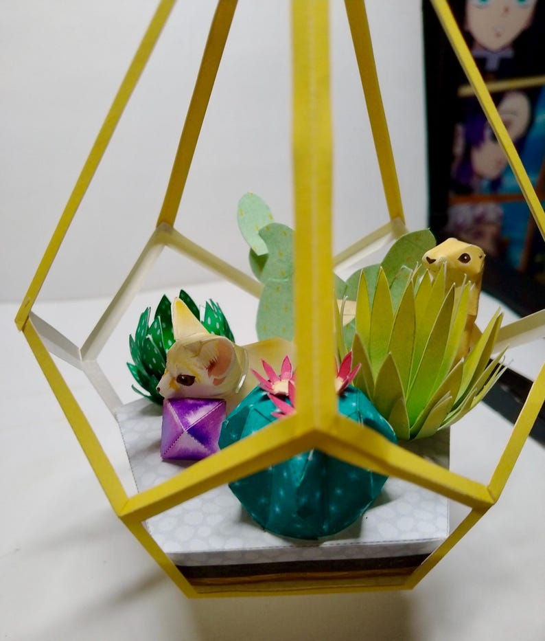 May include: A geometric yellow frame terrarium containing paper craft elements including a fennec fox, a small purple cube, a teal cactus with pink flowers, a light green succulent, and a spiky lime green plant on a white base with gray dots.