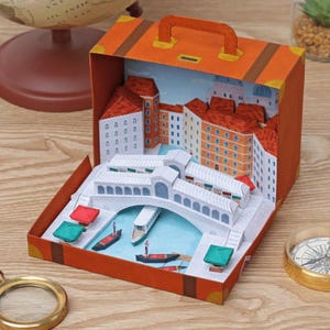 Puede incluir: Un diorama de papel en miniatura de Venecia, Italia, exhibido dentro de una maleta naranja. La escena incluye el Puente de Rialto, edificios con tejados rojos y góndolas en un canal azul. La maleta está abierta, revelando el arte en papel detallado.