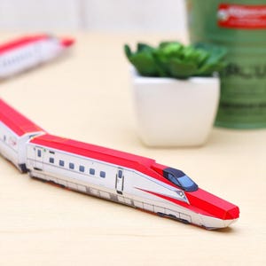 Puede incluir: Un modelo de tren detallado con una combinación de colores rojo y blanco. El tren presenta un diseño aerodinámico con un techo rojo y un cuerpo blanco. Las ventanas son visibles, y el tren parece estar hecho de papel o cartón.