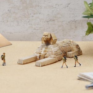 May include: A miniature Sphinx statue with a detailed carved design, in shades of beige and tan. Small figurines of people with backpacks are positioned nearby, suggesting a travel or historical theme. The scene is set on a light brown surface.