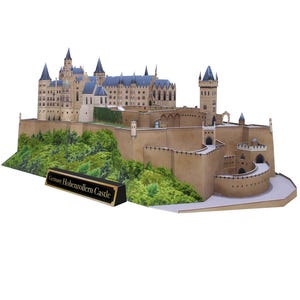 Op de afbeelding: Een gedetailleerd papieren model van kasteel Hohenzollern in Duitsland. Het kasteel is gemaakt van bruin papier en is omgeven door een grijze stenen muur. Het kasteel is omgeven door groene bomen en struiken. Het model staat op een witte achtergrond. De tekst "Duitsland Hohenzollern Kasteel" is gedrukt op een zwarte plaquette voor het model.
