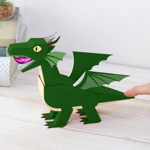 Op de afbeelding: Een groene papieren draak met een gele oog, roze tong en kleine hoorns. De draak heeft beweegbare vleugels en staat op een licht houten oppervlak. Het object heeft een fantasiethema.